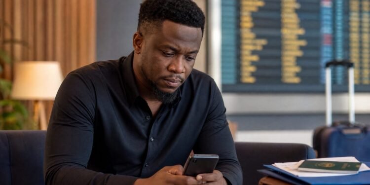 A Nigerian content creator sits thoughtfully with a smartphone near travel documents